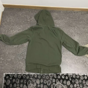 Grön huvtröja med dragkedja - Säljer en grön huvtröja med dragkedja och fickor framtill. Perfekt för en avslappnad stil. Huvan har vita snören för justering.