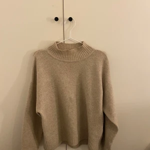 Beige stickad tröja - Mysig beige stickad tröja med hög krage och långa ärmar. Perfekt för kyliga dagar och ger en avslappnad look. Ribbstickade detaljer vid krage, ärmslut och nederkant.