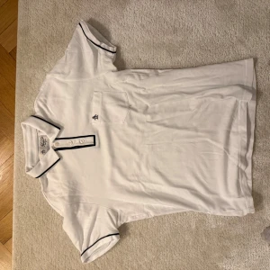 Vit pikétröja med svarta detaljer - Snygg vit pikétröja från Penguin med svarta detaljer på krage och ärmslut. Tröjan har en knappslå med tre knappar och en liten broderad logga på bröstfickan. Perfekt för en stilren och avslappnad look. Storlek M. Mycket bra skick