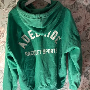 Grön hoodie med dragkedja - Säljer en grön hoodie med texten 'Adelaide Racquet Sports' på ryggen. Den har en dragkedja framtill och en mysig huva med snören. Perfekt för en avslappnad stil.