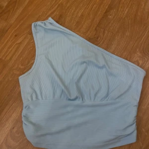 Ljusblå oneshoulder topp - Snygg ljusblå oneshoulder topp med ribbad textur. Perfekt för en stilren look. Toppen har en asymmetrisk design som ger en unik touch.