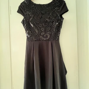 Svart klänning med broderade detaljer - Säljer en elegant svart klänning med vackra broderade detaljer på framsidan. Klänningen har en öppen rygg med knytband och korta ärmar. Perfekt för festliga tillfällen!