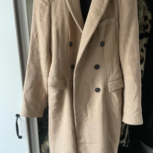 Beige kappa från Zara Woman - Säljer en stilren beige kappa från Zara Woman i storlek S. Den har en klassisk dubbelknäppt design med svarta knappar och långa ärmar. Perfekt för höst och vår, och ger en elegant touch till vilken outfit som helst. Passar både till vardags och festliga tillfällen.