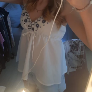 Vit blommig topp med knytband - Säljer en superfin vit topp med blommigt mönster och knytband. Toppen har en v-ringad design med broderade blommor i mörkblått. Perfekt för sommardagar eller en avslappnad festkväll. Passar bra till jeans eller kjol!