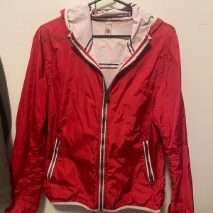 Röd vindjacka från Colmar - Säljer en snygg röd vindjacka från Colmar. Den har en dragkedja framtill och två praktiska fickor med dragkedjor. Jackan har en vit insida med ränder i blått och rött, vilket ger en fräsch och clean look. Perfekt för vår och höst!