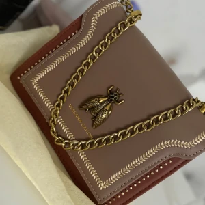 Brun axelväska med guldkedja och insektdetalj - Snygg brun axelväska från Vessina Gahon med en cool guldkedja och en unik insektdetalj på framsidan. Väskan har ett elegant broderat mönster och är perfekt för både vardag och fest. Den har en praktisk dragkedja inuti för säker förvaring. Helt ny