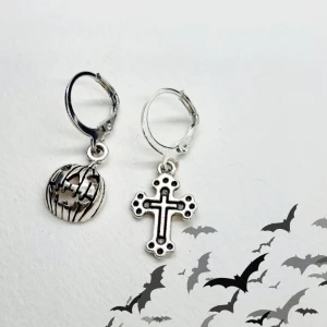 Silverörhängen med kors och pumpa - Säljer ett par unika örhängen med olika motiv. Det ena örhänget har en detaljerad korsdesign medan det andra har en cool pumpaform. Perfekta för att ge din outfit en edgy touch, speciellt under höstsäsongen eller till Halloween! 🦇Material-metall