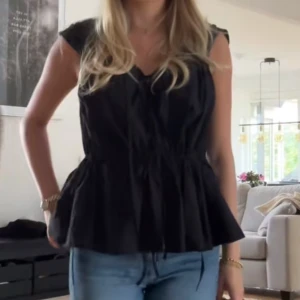 Svart ärmlös topp med knytning - Säljer en snygg svart ärmlös topp med knytning framtill. Toppen har en peplum-design som ger en fin siluett och är perfekt för både vardag och fest. Materialet känns lätt och luftigt, vilket gör den bekväm att bära hela dagen.