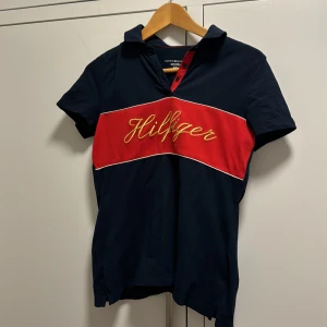 Mörkblå t-shirt från Tommy Hilfiger - Säljer en snygg mörkblå piké från Tommy Hilfiger. Den har en röd rand över bröstet med 'Hilfiger' broderat i gult. T-shirten är kortärmad och har en klassisk krage med knappar. Perfekt för en casual look!