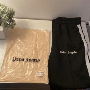 Svarta byxor från Palm Angels - Säljer ett par svarta byxor från Palm Angels i storlek M. Byxorna har en vit rand längs sidorna och elastisk midja. De är tillverkade av 100% bomull och polyester. Perfekta för en avslappnad och stilren look.