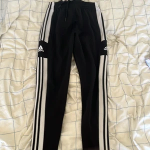 Svarta Adidas mjukisbyxor med vita ränder - Säljer ett par svarta Adidas mjukisbyxor i storlek M. Byxorna har de klassiska tre vita ränderna längs sidorna och Adidas-loggan på låren. De har en elastisk midja med snörning för justerbar passform och dragkedjor vid bensluten. Perfekta för träning eller en avslappnad dag hemma.