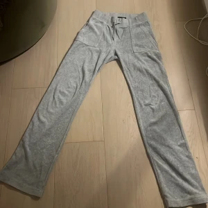 Grå mjukisbyxor från Juicy Couture - Säljer ett par supermysiga grå mjukisbyxor från Juicy Couture i storlek XS. Byxorna har en bekväm passform med snörning i midjan och stora fickor fram. Perfekta för en chill dag hemma eller en snabb tur till affären. De är i bra skick och redo för en ny ägare!