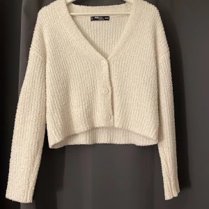 Vit croppad kofta från FB Sister - Supermjuk och fluffig vit kofta från FB Sister. Croppad modell med v-ringning och tre knappar framtill. Långärmad och ribbad struktur som ger en mysig känsla. Perfekt att slänga på sig över en topp när det är kyligt.