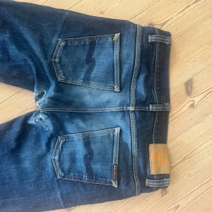 Mörkblå Nudie Jeans Skinny - Säljer ett par mörkblå skinny jeans från Nudie Jeans med klassiska kontrastsömmar och snygga slitningar. Jeansen har fem fickor, orange detaljer och Nudie-logga på bakfickan. Perfekt passform med smal siluett och stretchigt jeanstyg. Storleken är 33 i midjan och jag har själv 34, dom kommer sitta perfekt på dig som har mellan 31-32-33