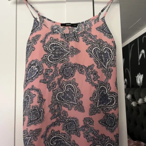 Rosa mönstrad klänning från Oasis - Supersöt rosa linne från Oasis med tunna axelband och blått paisleymönster med hjärtan. Lätt och flowy modell som är perfekt för varma dagar. Snygg till sneakers eller sandaler och enkel att styla med accessoarer.