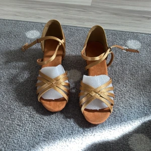 Guldiga (latinodans) klackskor med låg klack - Snygga klackskor i guldig satin med korsade remmar framtill och öppen tå. Skorna har en låg klack och justerbar rem runt vristen. Perfekta för dig som vill sticka ut med en elegant och glansig look.