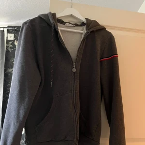 Mörkgrå hoodie från Moncler med huva - Snygg mörkgrå hoodie från Moncler med dragkedja, huva och fickor framtill. Ärmen har en röd, vit och svart rand samt Moncler-logga. Perfekt för dig som gillar stilrena och sportiga plagg. Tillverkad i mjuk bomullsmix för extra komfort. Den är köpt från Von greven. Storlek L men passar S/xs