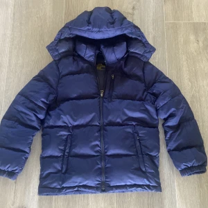 Mörkblå pufferjacka från Ralph Lauren - Säljer en mörkblå pufferjacka från Ralph Lauren Performance med huva och broderad logga på bröstet. Jackan har dragkedja framtill, två sidofickor med dragkedja och elastiska muddar vid ärmslut. Perfekt för kalla dagar och riktigt snygg stil.  Logan där inne har lossnat lite i tvättet men det är ingen fara, på sista bilden ser att tyget har börjat lossna litegrann vid armen men det syns inte om man inte kollar riktigt noga. Annars är det en väldigt snygg jacka 