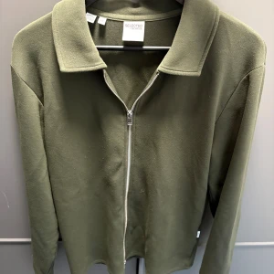 Olivgrön ziptröja från Selected Homme - Olivgrön långärmad tröja från Selected Homme med hel dragkedja framtill och klassisk krage. Tröjan har en clean look och är tillverkad i mjukt material som känns skönt mot huden. Perfekt för lager-på-lager-stil.