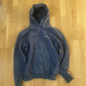 Mörk grön hoodie från Champion - Mysig mörkgrön hoodie från Champion med broderad logga på bröstet och klassisk känguruficka framtill. Tröjan har huva med dragsko och långa ärmar med ribbade muddar. Passar bra  för chill dagar eller när du vill ha en avslappnad stil. Stilren, fullt äkta!