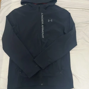 Svart vindjacka från Under Armour - Svart vindjacka från Under Armour med huva och hel dragkedja framtill. Jackan har logga och text i silver på bröstet, samt ventilerande meshfoder på insidan. Snygg och sportig design med praktiska fickor med dragkedja.