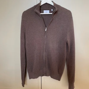 Brun full zip från Bläck - Säljer en stilren brun kofta från Bläck i storlek S. Koftan är stickad, har hel dragkedja framtill och en hög krage. Perfekt för lager-på-lager och passar till många olika outfits. Enkel och klassisk design som är lätt att matcha.