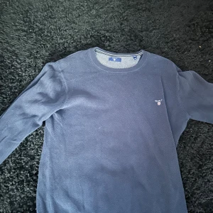 Mörkblå stickad tröja från GANT - Säljer en mörkblå stickad tröja från GANT med diskret logga broderad på bröstet. Tröjan har rund halsringning och långa ärmar. Perfekt för dig som gillar klassisk och stilren design.