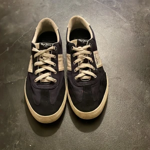 Mörkblå sneakers från Pepe Jeans - Snygga mörkblå sneakers från Pepe Jeans med vita detaljer och snörning. Skorna har en klassisk låg profil, vit sula och rund tå. Perfekta för dig som gillar en stilren och sportig look. Materialet är textil och syntet. 