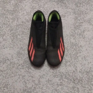 Adidas Crazyfast - Säljer ett par svarta adidas Crazyfast  med orangeröda ränder på sidorna och gröna detaljer vid hälen. Skorna har en strömlinjeformad design utan snörning och är tillverkade i syntetmaterial för bästa grepp och känsla på planen.