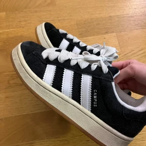 Adidas Campus sneakers svart/vit - Svarta Adidas Campus sneakers i mocka med klassiska vita ränder och breda vita skosnören. Låg modell med vit sula och Adidas-logga på hälen. Snygg retrostil som funkar till det mesta.