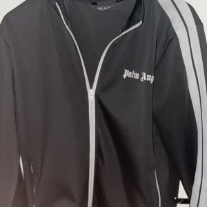 Palm Angels svart track jacket - Svart track jacket från Palm Angels med vita ränder längs ärmarna och vit dragkedja. Jackan har logga på bröstet och är tillverkad i polyester. Perfekt för en sportig och trendig streetwear-look.