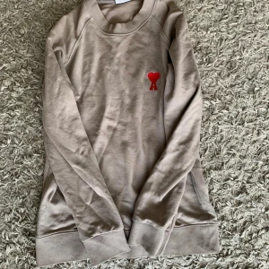 Beige sweatshirt från Ami Paris - Säljer en beige sweatshirt från Ami Paris med röd broderad hjärtlogga på bröstet. Tröjan är i mjuk bomull, har rund hals och långa ärmar. Perfekt för dig som gillar stilrena och trendiga plagg med en twist.