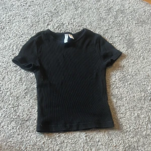 Svart T-shirt - Säljer denna svarta T-shirt i storlek xs då den inte används längre. Använts fåtal gånger. Den är ribbad och inte så lång.
