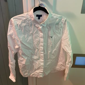 Vit skjorta från Gant (Barn) - Klassisk vit skjorta från Gant med broderad logga på bröstet. Skjortan har lång ärm, knappar framtill och en stilren krage. Perfekt för en clean och tidlös look. Tillverkad i bomull för en skön känsla hela dagen.
