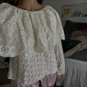 Vit spetsblus med volang - Superfin vit blus i spets med stora volanger över axlarna och långärmad modell. Blusen har ett vackert broderat mönster och är lätt transparent. Perfekt för dig som gillar romantisk och bohemisk stil.