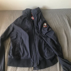 Mörkblå vindjacka från Parajumpers - Snygg mörkblå vindjacka från Parajumpers med dragkedja framtill och ribbade muddar. Jackan har en klassisk patch på ärmen och två snedställda fickor. Perfekt för dig som vill ha en stilren och sportig look.