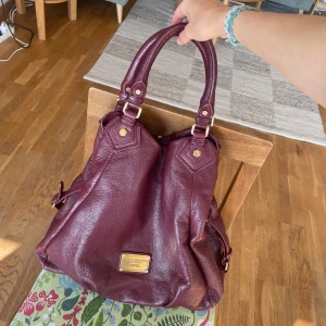 Burgundy handväska Marc by Marc Jacobs - Supersnygg vinröd handväska från Marc by Marc Jacobs i äkta skinn med guldfärgade metalldetaljer och dubbla handtag. Den har en cool innerfodring med texten ”Marc Jacobs” Perfekt storlek för skola/jobb då den rymmer laptop och mappar! I helt perfekt skick, skriv vid frågor😊