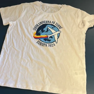 Vit t-shirt Järnkaminerna Europa 2024 - Vit t-shirt med tryck av ett flygplan och texten 'Järnkaminerna på turné Europa 2024' över en blå jordglob. Färgglad svans i rött, gult och blått. Klassisk passform och rund hals. Perfekt för dig som vill visa support på resan!