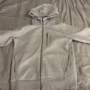 Grå hoodie från Sail Racing med dragkedja - Snygg ljusgrå hoodie från Sail Racing med hel dragkedja och huva med snörning. Tröjan har en bröstficka med dragkedja och två sidofickor. Perfekt för kyliga dagar och har en clean, sportig look.