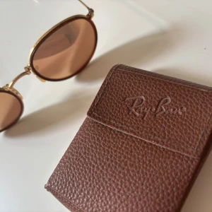 Runda solglasögon Ray-Ban guld - Säljer ett par Ray-Ban solglasögon med rund form och guldfärgade metallbågar. Glasen är bruna med en varm ton och bågarna har snygga detaljer. Kommer med ett brunt Ray-Ban fodral i skinnimitation. Perfekt för dig som gillar retrostil och klassisk design.