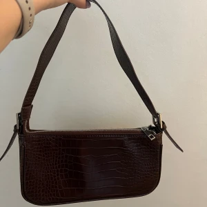 Brun axelväska i croco-mönster - Snygg mörkbrun axelväska med croco-mönstrad yta och silvrig dragkedja. Väskan har en bred axelrem🤎Supersnygg nu till hösten!