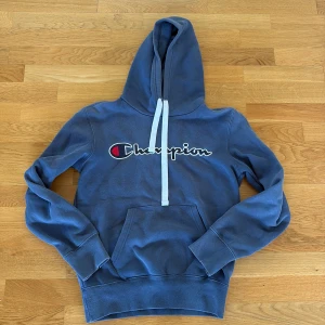 Blå hoodie från Champion - Säljer en blå hoodie från Champion. Tröjan har huva med vita snören, broderad Champion-logga på bröstet och en klassisk känguruficka framtill. Smal modell. Storlek S. Hel och ren.