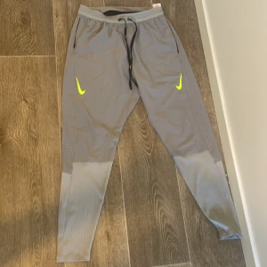 Grå träningsbyxor från Nike - Snygga grå träningsbyxor från Nike med svarta snören i midjan och gula/gröna loggor på båda benen. Byxorna har ribbade detaljer nedtill och är tillverkade i ett lätt och stretchigt material som passar perfekt för sport och aktivitet.
