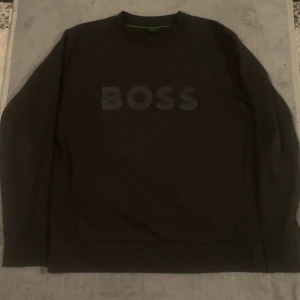 Mörkblå sweatshirt från BOSS, storlek M - Mörkblå sweatshirt från BOSS i storlek M med klassisk rund hals och stort BOSS-tryck framtill. Tröjan har långa ärmar och en clean look med diskreta gröna detaljer på insidan av kragen. Perfekt för en stilren och avslappnad outfit.
