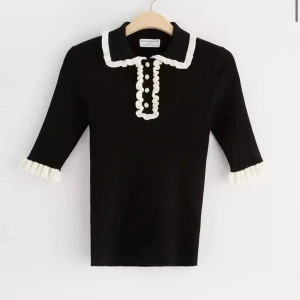 Svart pikétröja med volang från Holly & Whyte - Supersöt svart pikétröja från Holly & Whyte med krämvita volangdetaljer vid krage, ärmslut och knappslå. Tröjan har pärlknappar och är ribbad i materialet. ANDVÄND EN GÅNG fint sick är stretchig. finns extra knapp med!💞