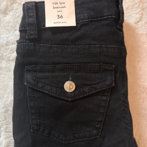 Svarta Y2K low bootcut jeans - Säljer ett par svarta Y2K-inspirerade low waist bootcut jeans från Gina Tricot. Byxorna har klassiska fickor fram och bak, samt en snygg knappdetalj på bakfickan. Aldrig använda och är i short lenght 