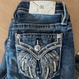 Miss Me jeans med broderade fickor - Snygga blå jeans från Miss Me med broderade bakfickor och paljettdetaljer. Jeansen har vita kontrastsömmar, klassisk femficksmodell och en bootcut passform. Perfekta för dig som gillar statement-jeans med coola detaljer.