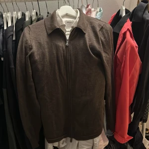 Brun stickad full zip från J.Lindeberg - Snygg brun stickad jacka från J.Lindeberg i storlek S. Jackan har dragkedja framtill, klassisk krage och lång ärm. Perfekt för dig som gillar stilrena och enkla plagg med en clean look. Merinoull, jätte bra skick! Sitter perfekt på mig som är 1,80 och 60 kg