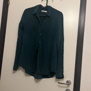 Mörkgrön skjorta från Gina Tricot - Säljer en mörkgrön skjorta från Gina Tricot i storlek 34. Skjortan har klassisk krage, knäppning framtill och långa ärmar. Materialet är mjukt och har en lätt glansig finish som ger en snygg look. Perfekt att styla med jeans eller kjol.
