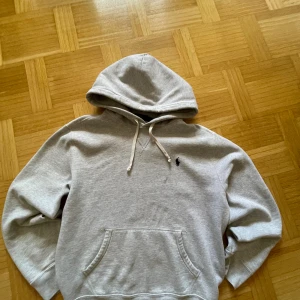 Grå hoodie från Polo Ralph Lauren - Snygg ljusgrå hoodie från Polo Ralph Lauren med klassisk broderad logga på bröstet. Tröjan har huva med snörning, känguruficka och är tillverkad i mjuk bomullsmix. Perfekt för en chill och stilren look. Tröjan är i storlek M men passar mer som S